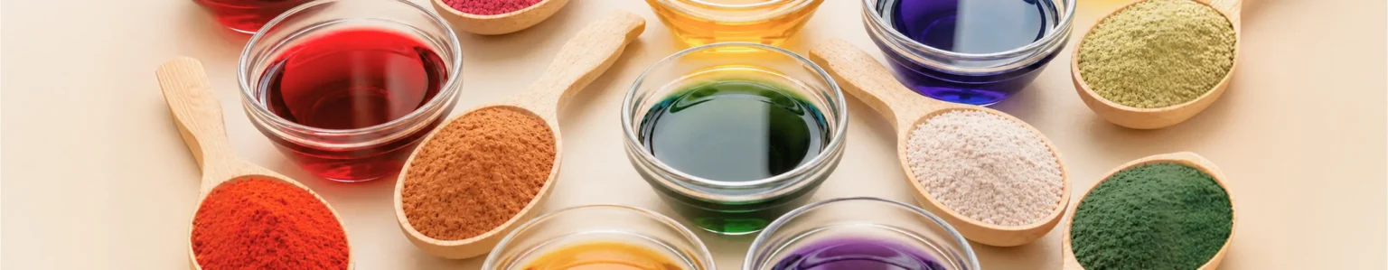 Variedade de corantes naturais Biocarmim em líquido e pó, nas cores vermelho, amarelo, azul, verde e roxo em tigelas e colheres de madeira